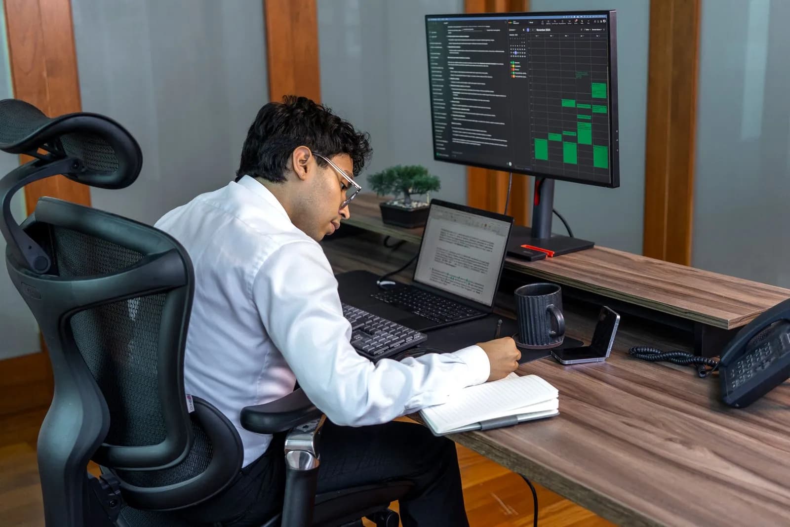 Un collaborateur travaille à son bureau entre un ordinateur portable et un écran affichant un tableur.